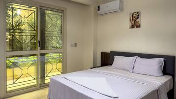 A hotel room with a bed, a window with a view of greenery, and an air conditioning unit on the wall.
