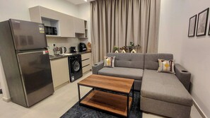 A compact living space with a kitchenette, a grey sofa set, a wooden coffee table, and a large window with curtains. - 832 . Modern 1BR Apartment | Near AUC (New Cairo City)