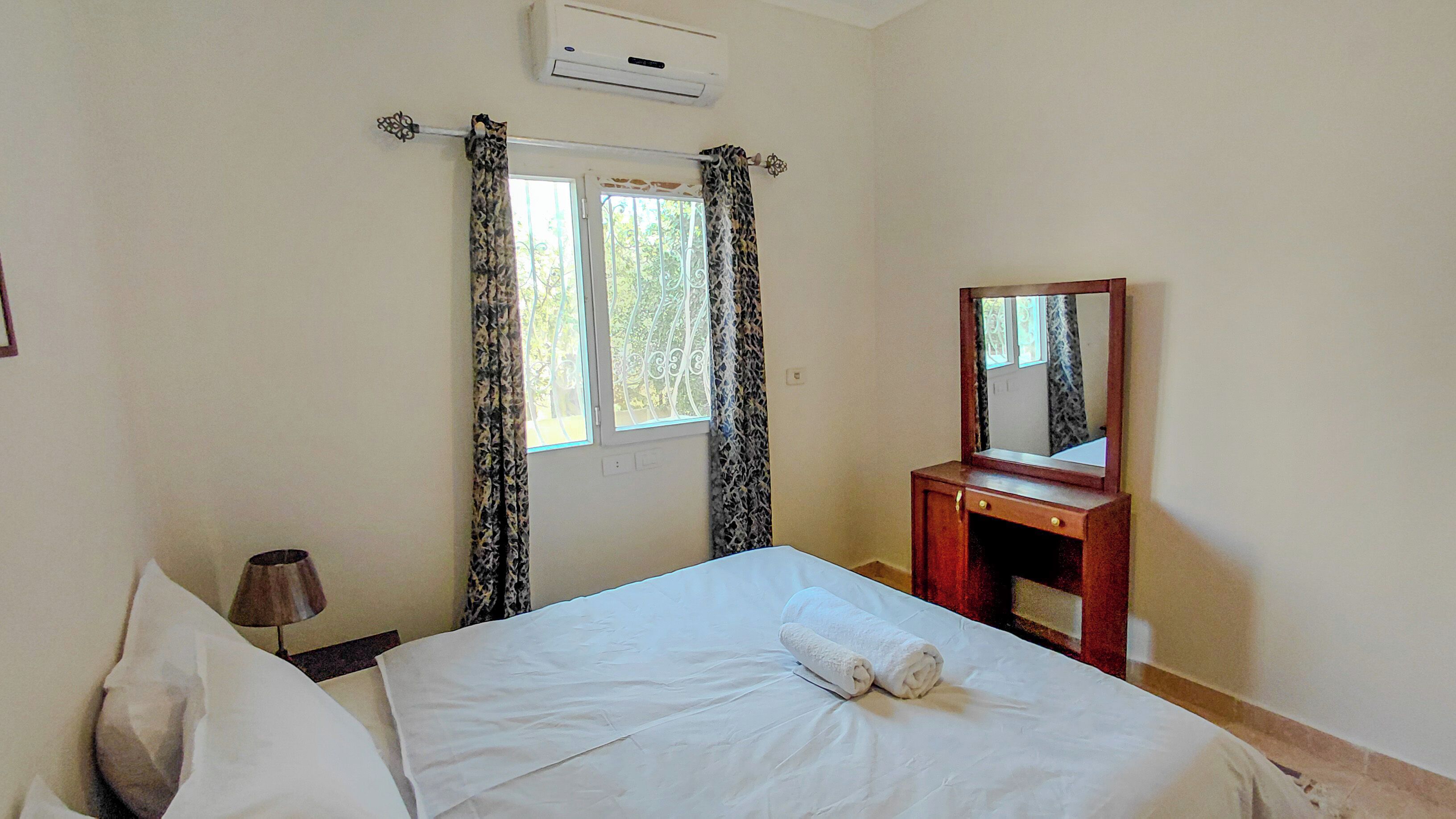 A bedroom with a bed, a bedside table, a mirror, and a window with curtains.