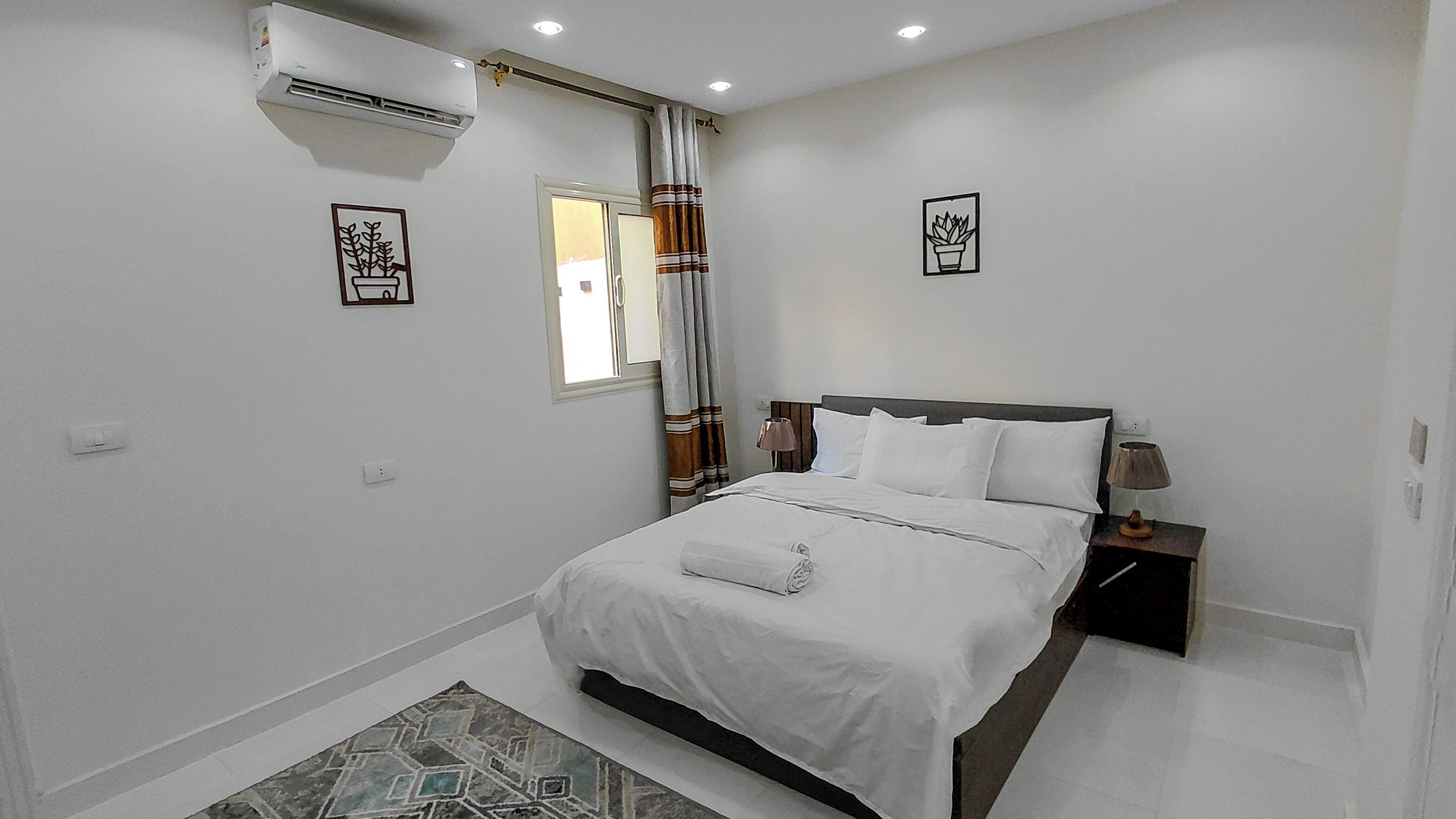 A bedroom with a bed, bedside table, lamp, air conditioning unit, and a window with curtains.