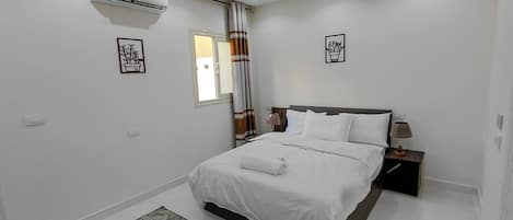 A bedroom with a bed, bedside table, lamp, air conditioning unit, and a window with curtains.