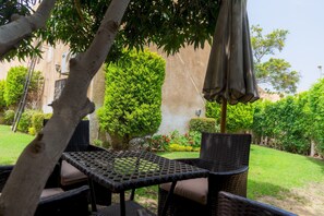 Un espacio al aire libre con mobiliario de mimbre y una mesa, rodeado de follaje y un edificio al fondo.