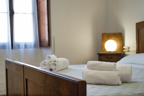 A neatly made bed with rolled white towels, a bedside table with a lamp, and a window with curtains.
