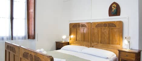 Una cama de madera con cabecera, dos mesitas de noche, una lámpara fijada a la pared y una ventana con cortinas.