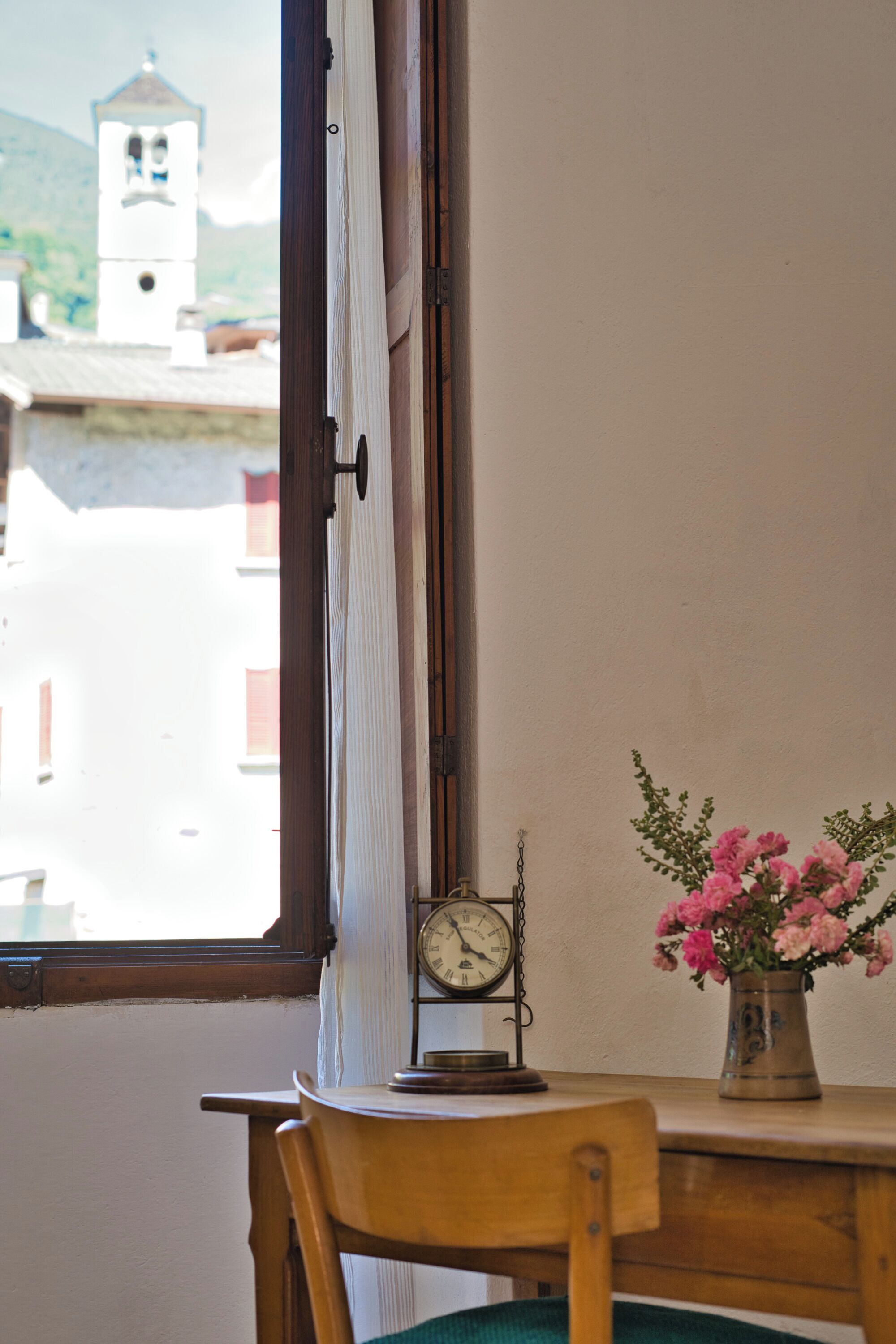 A wooden chair, a table with a clock and a vase of flowers, and a window with a view of a building and a bell tower.