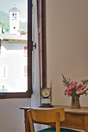 Ein Holzstuhl, ein Tisch mit einer Uhr und einer Blumenvase, sowie ein Fenster mit Blick auf ein Gebäude und einen Glockenturm.