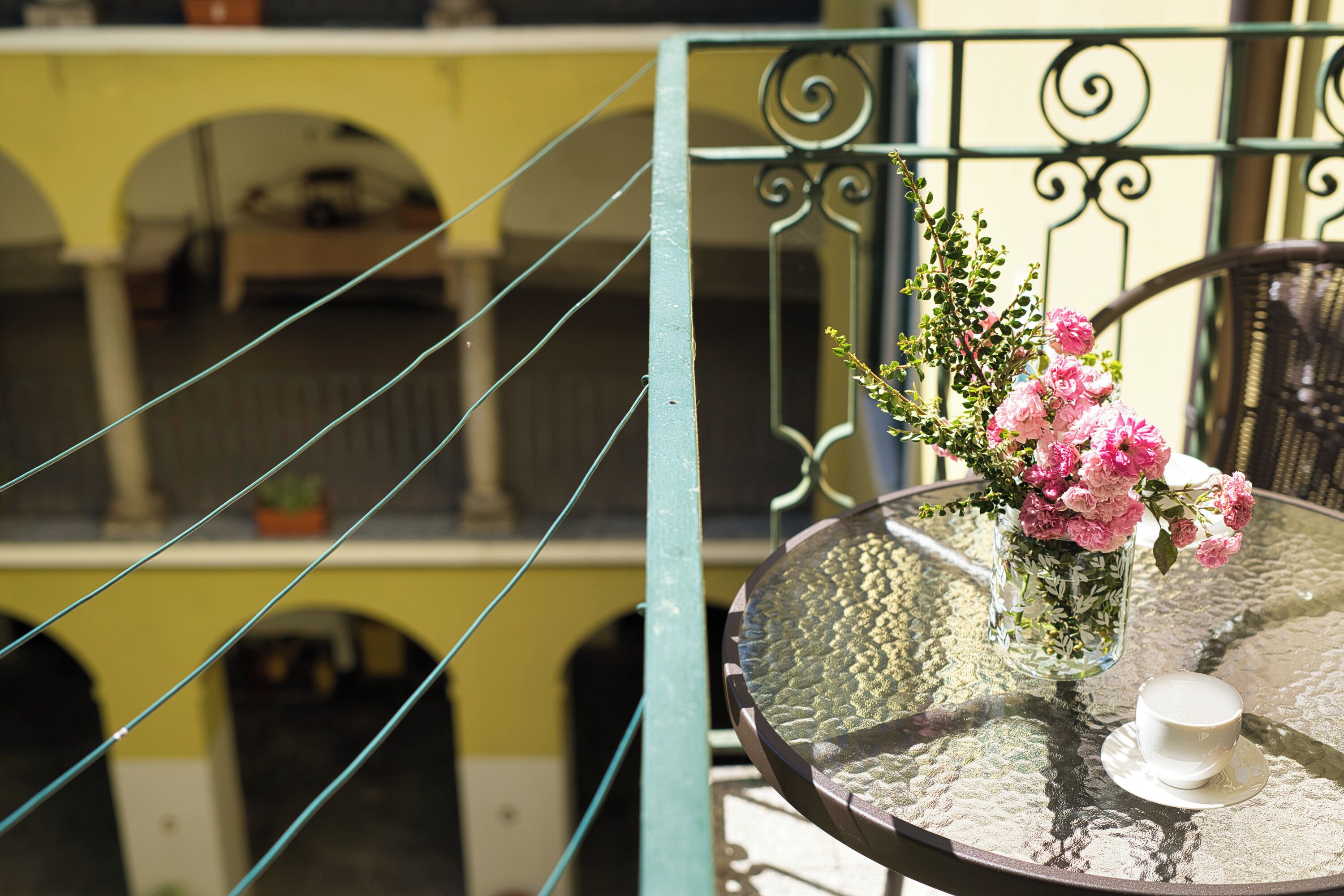 A balcony with a metal table, a glass vase with pink flowers, and a white cup.