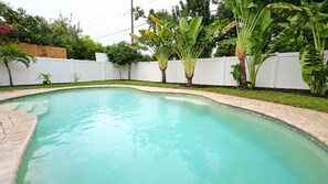 Ein Schwimmbecken mit geschwungener Kante, umgeben von einem weißen Zaun und üppiger Bepflanzung.