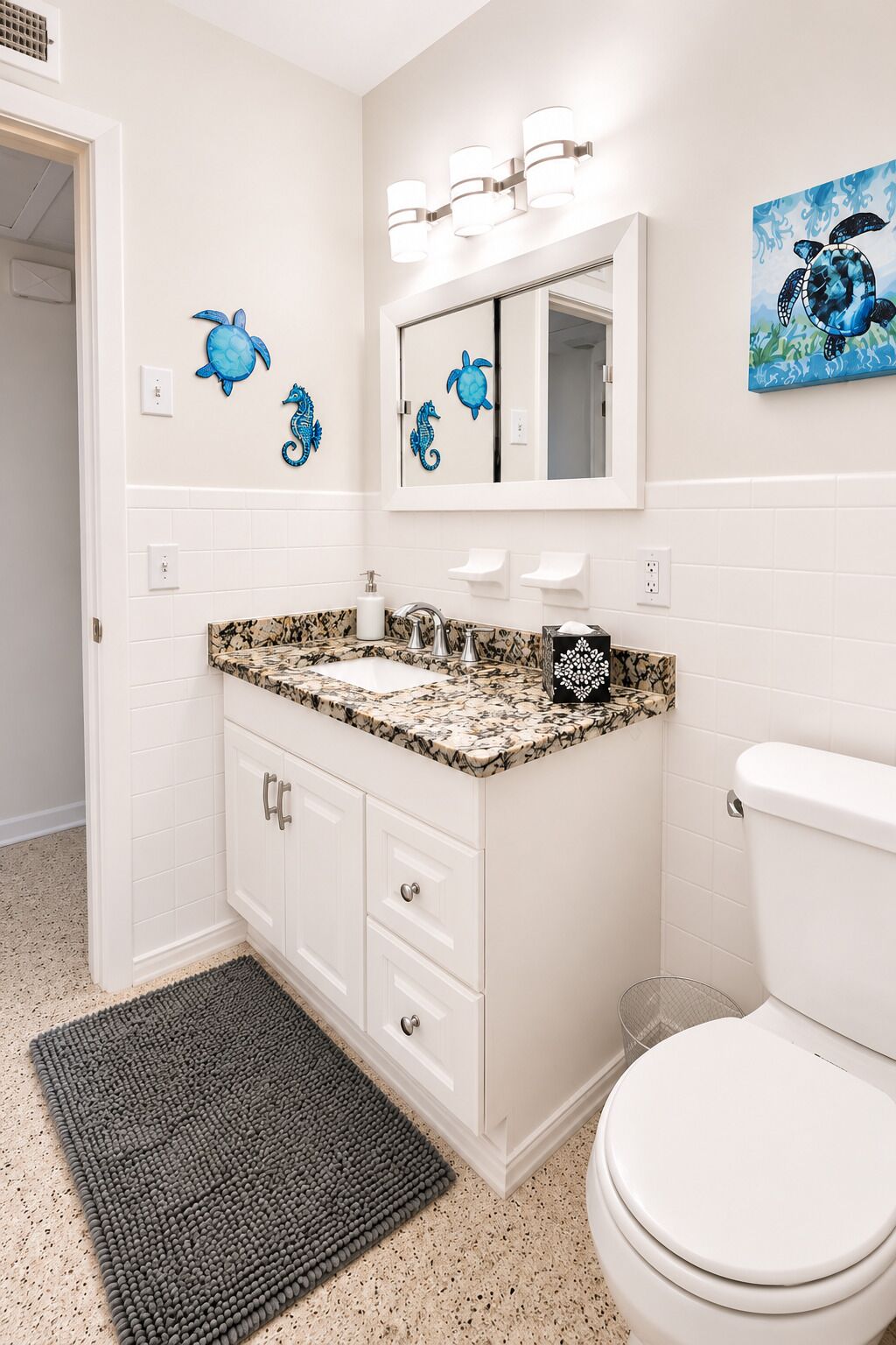 A bathroom with a white vanity, a sink with a speckled countertop, a mirror with wall-mounted lights, and a toilet.