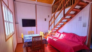 Una habitació amb sòl de fusta, un llit amb llençol vermell, una taula de fusta amb cadires, un televisor fixat a la paret i una escala.