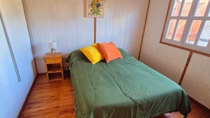 Habitació petita amb un llit individual, un terra de fusta, un estovat verd, una petita taula de nit amb una làmpada i una finestra amb vistes.