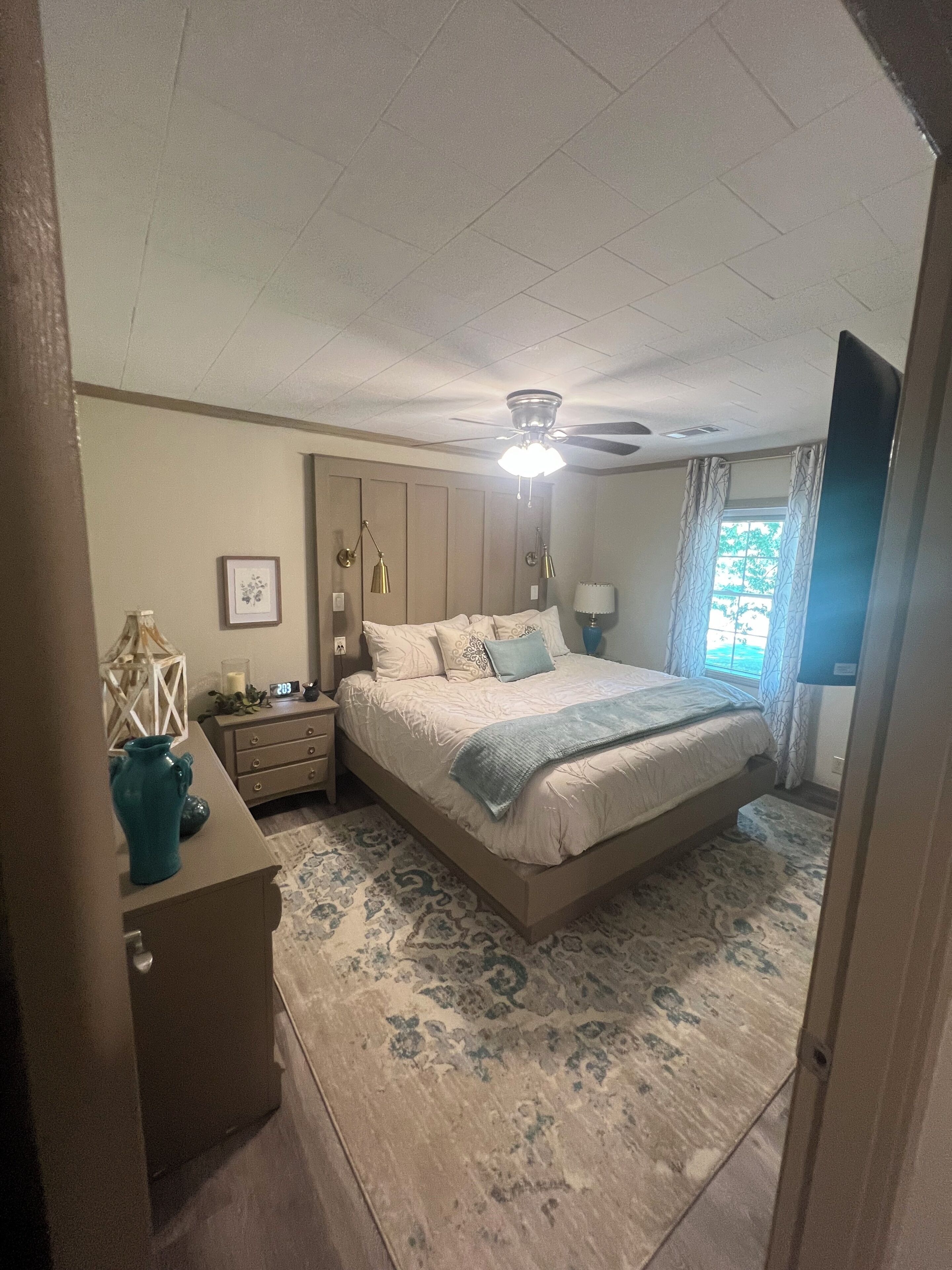 Un dormitorio con cama, mesitas de noche, lámpara de noche, ventilador de techo y ventana con cortinas.