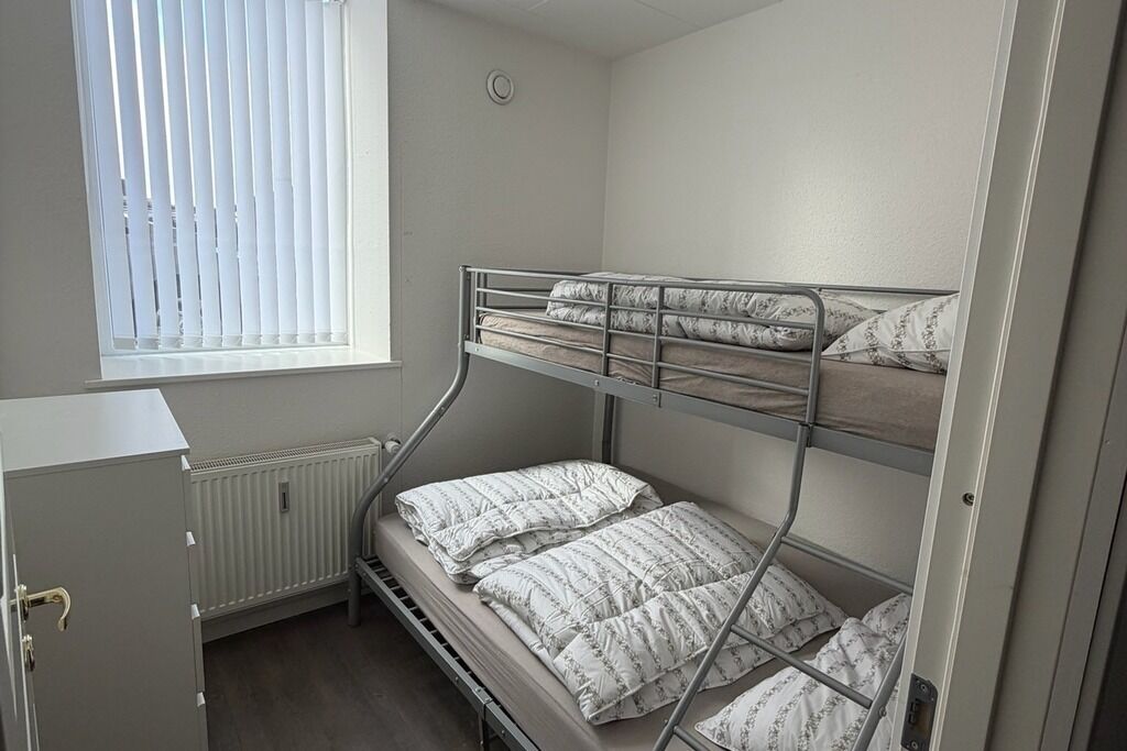 Une chambre avec un lit superposé, un radiateur, une fenêtre avec des stores et une porte.