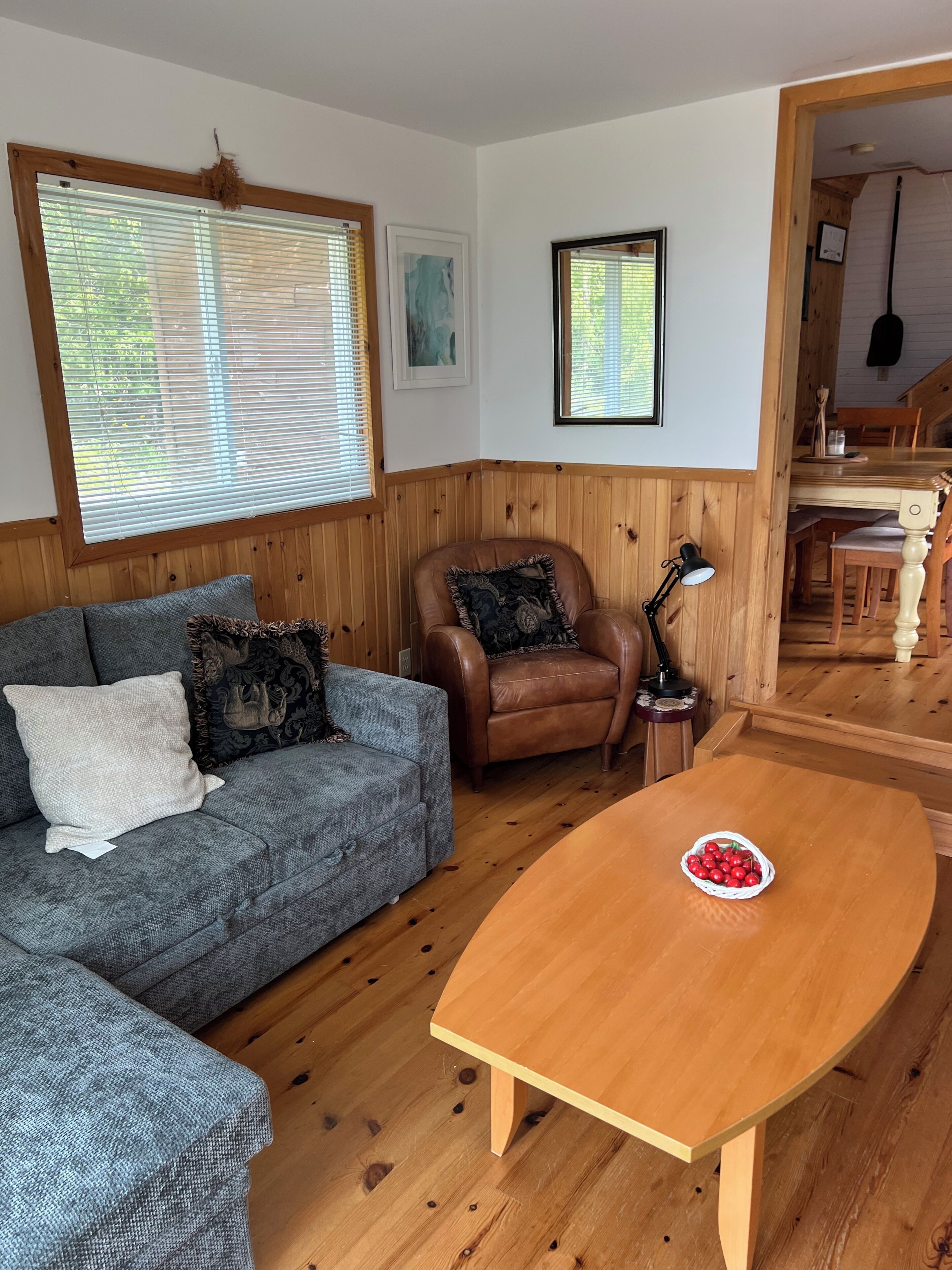 A cozy living room with a grey sofa, a brown leather chair, a wooden coffee table with a basket of cherries, and wooden paneled walls.