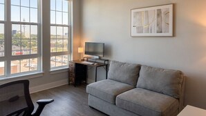Una stanza con un divano grigio, una scrivania con un computer e una TV. Sono presenti ampie finestre con vista su edifici e un parcheggio.