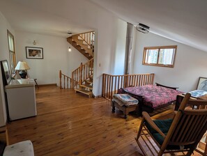 A room with a wooden floor, a bed with a patterned bedspread, a chair, a desk, a lamp, a picture frame, and a staircase.