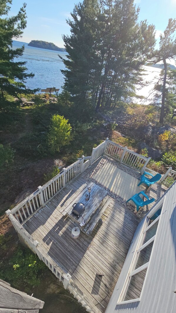 A wooden deck with a grill and chairs, overlooking a body of water and trees.