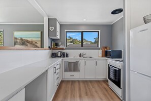 A modern kitchen with white cabinets, a stainless steel dishwasher, a built-in oven, and a window with a view of houses. - Calden Cottage - Ngunguru Holiday Home (Whangarei)