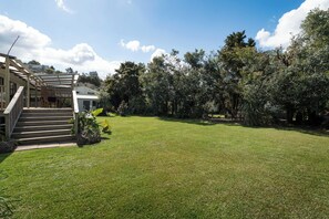 Um imóvel residencial com gramado bem cuidado, uma escadaria de madeira que leva a um alpendre coberto, e diversas árvores e arbustos.