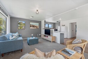 A modern living room with a sofa, ottoman, and a dining area with a TV and kitchen counter. - Calden Cottage - Ngunguru Holiday Home (Whangarei)
