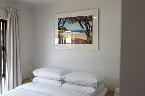 A bedroom with a bed, white pillows, a framed picture of a beach scene, and a window with curtains. - Calden Cottage - Ngunguru Holiday Home (Whangarei)