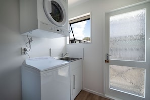 A compact laundry room with a washing machine, a sink, and a window. - Calden Cottage - Ngunguru Holiday Home (Whangarei)