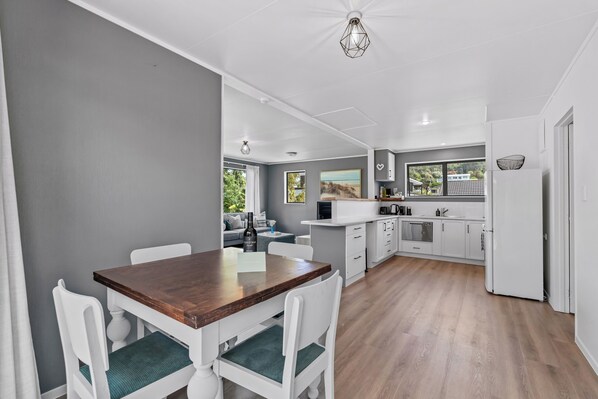 A modern kitchen with white cabinetry, a wooden dining table, and a view of the outdoors. - Calden Cottage - Ngunguru Holiday Home (Whangarei)