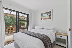 A bedroom with a bed, a nightstand, a framed picture, and a view of an outdoor deck. - Calden Cottage - Ngunguru Holiday Home (Whangarei)