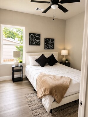 Ein Schlafzimmer mit Bett, Deckenventilator, Wandbild, Fenster und Nachttisch mit Lampe.