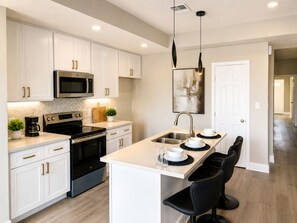 A modern kitchen with white cabinetry, a stainless steel microwave, a built-in oven, and a central island with bar stools. - Executive 3BR  Near Downtown, Port & LBJ Hospital (Houston)