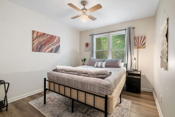 A bedroom with a bed, a ceiling fan, a window with curtains, a nightstand, and a wall-mounted lamp.