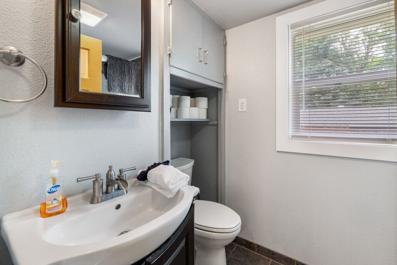 Une salle de bain avec un lavabo blanc, des toilettes, un miroir et une fenêtre avec des stores.