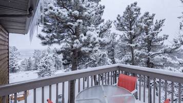 A snow-covered balcony with a table and chairs, overlooking a snowy landscape with trees.