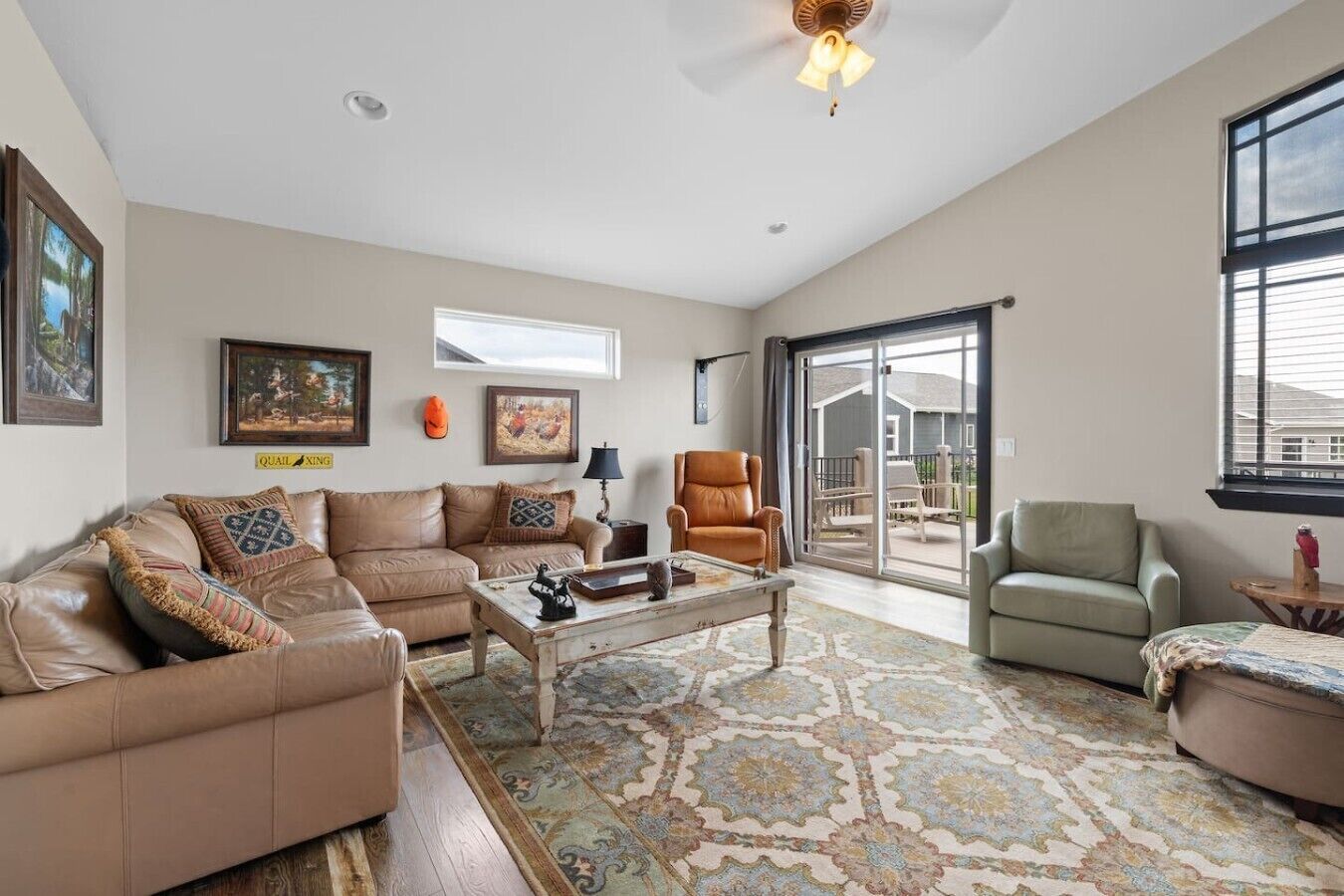 A spacious living room with a brown leather sofa, a coffee table, and a patterned rug. There is a sliding glass door leading to a balcony with outdoor furniture.