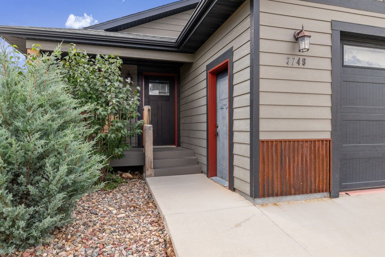 A residential building with a grey siding exterior, a red door, and a mailbox with the number 3749.