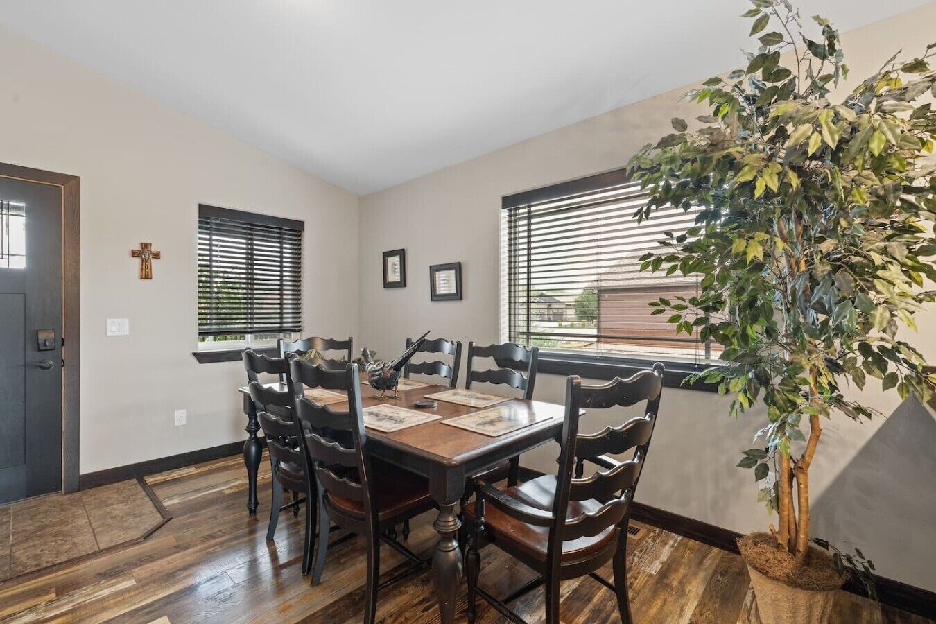A dining room with a wooden table and chairs, a cross on the wall, and a large plant.