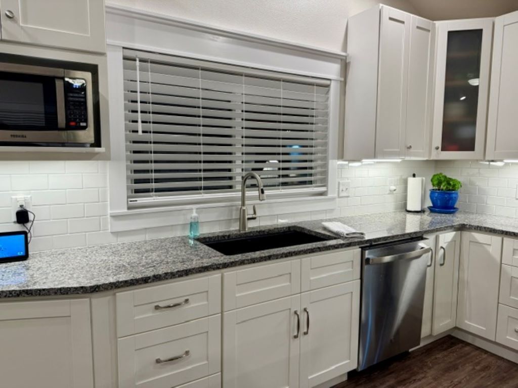 A kitchen with white cabinets, a stainless steel dishwasher, a microwave, and a window with blinds.