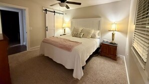 A bedroom with a bed, a ceiling fan, bedside lamps, a nightstand, and a door. - Two Tired Inn: Cozy, convenient, hot tub, fire pit, by trails, museums, downtown (Bentonville)