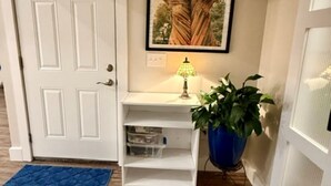 A hallway with a white door, a framed picture on the wall, a small white table with a lamp, a blue rug, and a potted plant. - Two Tired Inn: Cozy, convenient, hot tub, fire pit, by trails, museums, downtown (Bentonville)