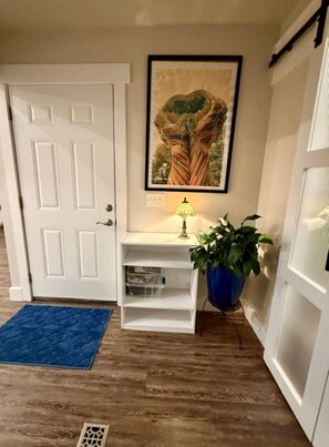Ein Flur mit einer weißen Tür, einem gerahmten Bild an der Wand, einem kleinen weißen Tisch mit einer Lampe, einem blauen Teppich und einer Topfpflanze.