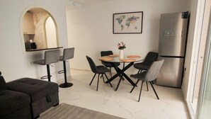Uma sala de estar moderna com um sofá seccional, uma área de jantar com mesa redonda e cadeiras, uma cozinha com geladeira e uma janela grande.