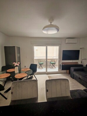 Ein Wohnzimmer mit Glastisch, Stühlen, einer Couch, einem Fernseher und einem Balkon mit Sitzgelegenheiten.