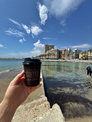 A hand holding a coffee cup with text, a clear sky, and a waterfront with buildings in the background. - La Casa Datskovi Beach Apartments (Torre Vieja)