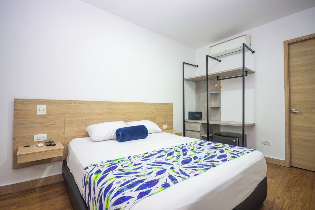 A hotel room with a bed, a bedside table with a phone and charger, a small desk with a radio, and a wardrobe with a mirror.