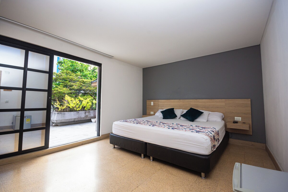 A modern bedroom with a large bed, a wooden headboard, and a sliding glass door leading to a balcony with greenery.