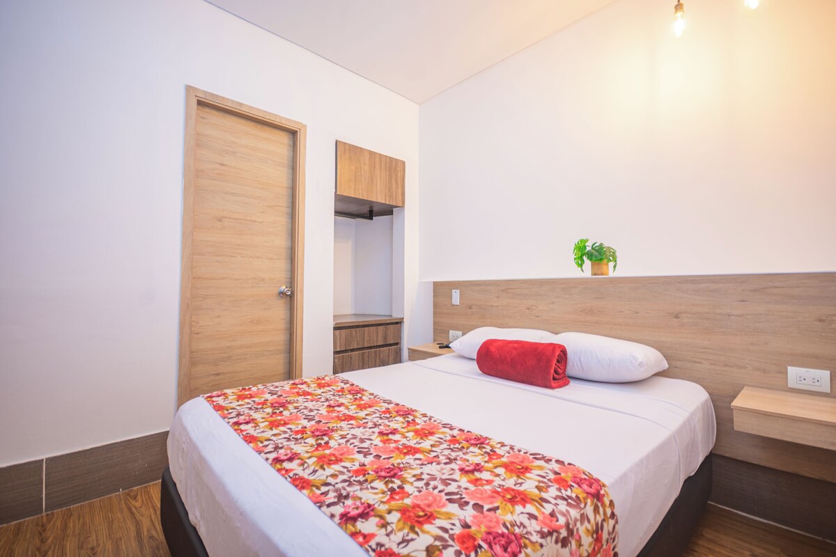 A hotel room with a bed, a wooden wardrobe, a small plant on the nightstand, and a door leading to a bathroom.