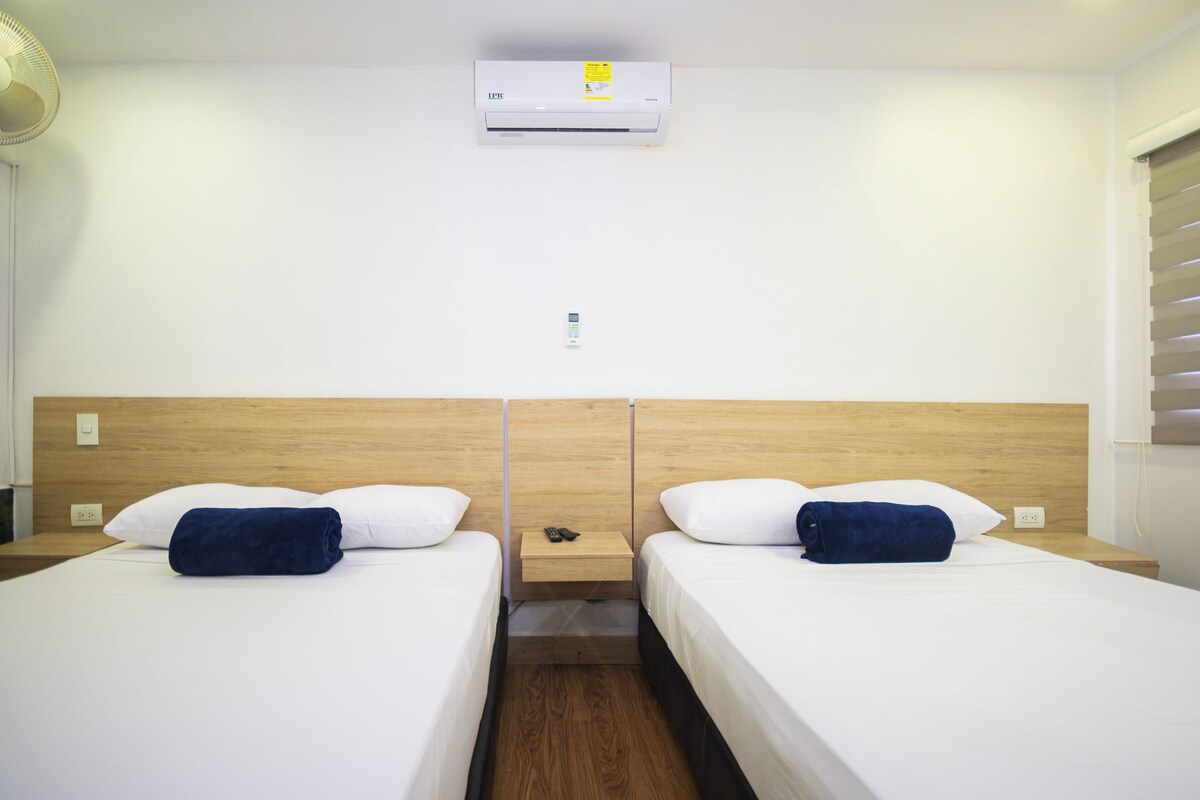 A hotel room with two beds, a wooden headboard, a wall-mounted air conditioner, and a bedside table with a remote control.