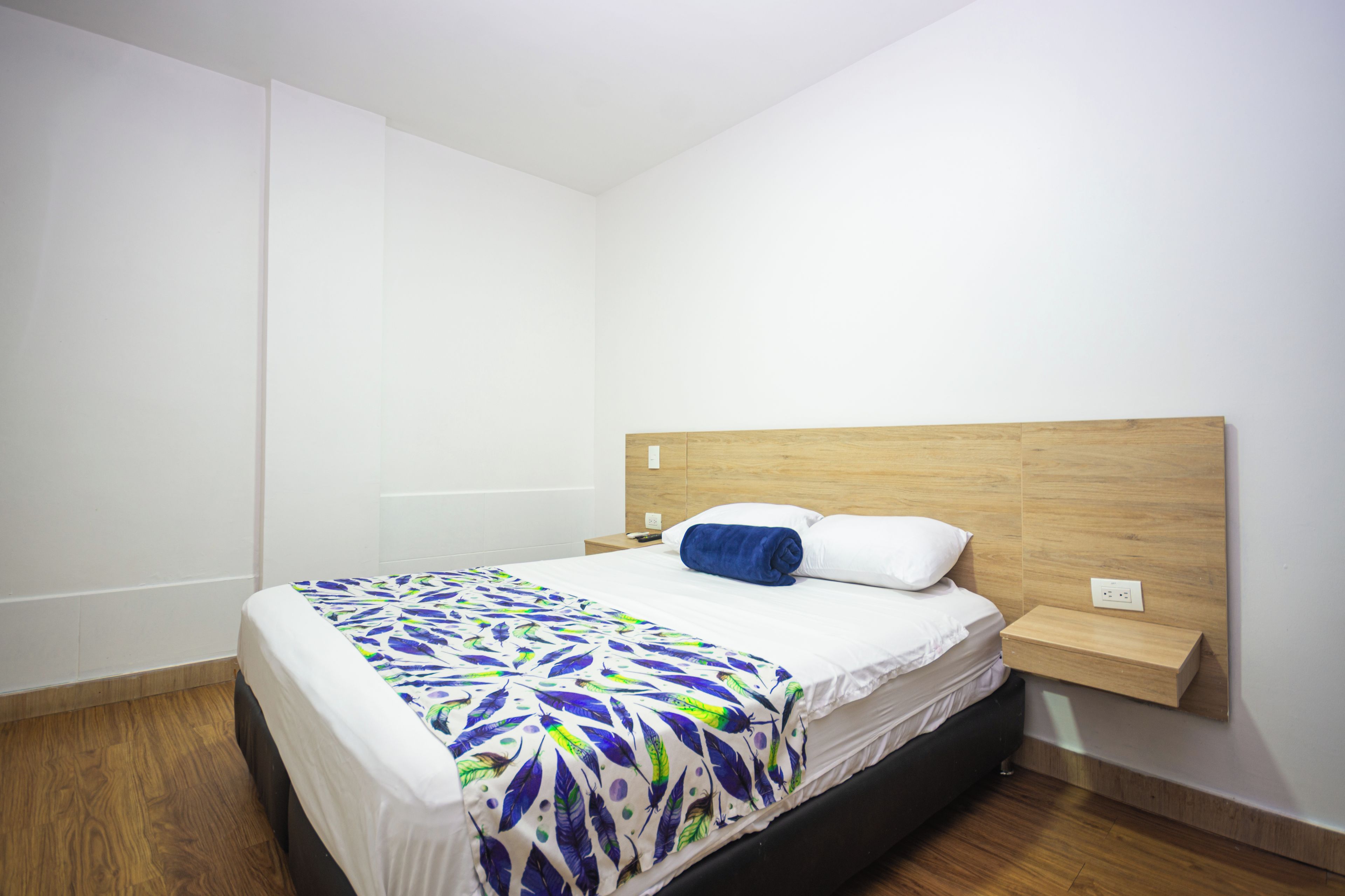 Ein Einzelbett mit einem gemusterten Überwurf, ein hölzernes Kopfteil, ein Nachttisch und eine Steckdose an der Wand.