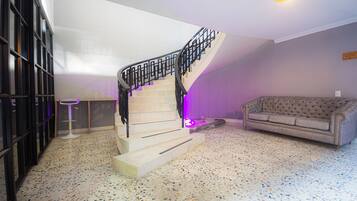 Un escalier avec une rampe noire menant à un palier, un canapé capitonné et un tabouret de bar dans une pièce dont le sol est recouvert d’un revêtement à motifs.