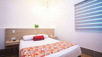 Une chambre d’hôtel avec une tête de lit en bois, un lit recouvert d’une couvre-lit à motifs floraux, un oreiller rouge, une table de chevet avec une télécommande et une petite plante posée sur la tête de lit.
